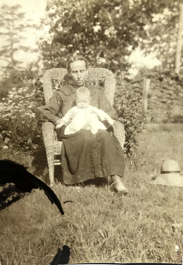A black-and-white photo of Mary Tinker Pyatt sitting outdoors in a wicker chair, holding her infant daughter, Cora, on her lap. Mary is dressed in a simple, long dark dress, her expression calm and reflective. Cora, dressed in a light-colored gown, rests on her mother's lap, facing the camera. The background shows a natural setting with trees and bushes, suggesting a quiet moment in a garden or rural yard. A hat rests on the grass beside them, adding to the peaceful and domestic atmosphere of the scene.