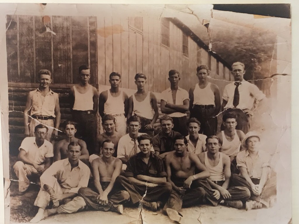 Vintage black and white photograph from the 1930s showing a group of seventeen men at what appears to be a Civilian Conservation Corps (CCC) camp. Most of the men are dressed in work attire—sleeveless undershirts and trousers. Delmer, distinguished by wearing a tie, stands in the back row on the right side. The group is posed in front of a large wooden building, possibly a barracks or workshop, with some men seated on the ground, others kneeling, and the rest standing. The photo captures a moment of camaraderie among the men, evident in their relaxed postures and casual attire, typical of a work camp environment from that era.