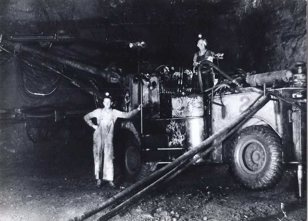 Black and white photograph of two miners at work in an underground setting, likely from the mid-20th century. On the left, a miner stands confidently, hands on hips, wearing a long, light-colored overcoat and a hard hat equipped with a headlamp. To the right, another miner is seated on a large, complex mining machine, also wearing a hard hat with a headlamp. The machine appears to be a type of drilling or excavation equipment, with visible gears and hydraulic components, set against the dark, rocky backdrop of the mine. The scene captures the gritty environment of lead mining, highlighting the robust machinery and the miners' readiness to tackle their demanding tasks.
