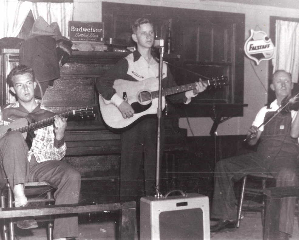 Black and white photograph of three men performing at a local event. From left to right: a young man seated, playing an acoustic guitar; a middle-aged man standing, playing an acoustic guitar; and an older man seated, playing the fiddle. They are on a simple wooden stage with music equipment around them, including a microphone and an amplifier. In the background, Budweiser beer advertisements are visible, suggesting the setting is a casual bar or community hall.