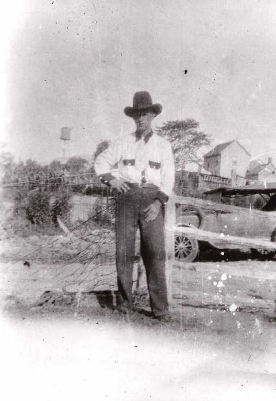 Black and white photograph of a man standing outdoors in a rural setting, circa early 20th century. The man is dressed in a white shirt with rolled-up sleeves, dark trousers, and a wide-brimmed hat, leaning casually against a wooden fence post. Behind him, a faded backdrop features old wooden buildings and a vintage car, suggesting a small-town or farming community. The image is slightly faded and speckled with age, adding a nostalgic quality to the scene.