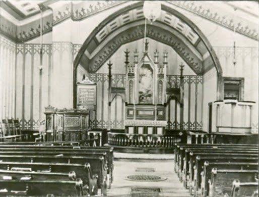 Black and white photograph of the interior of Lost Island Lutheran Church, showcasing an elegantly designed altar at the center of a semi-circular arch adorned with religious motifs. The church features traditional wooden pews arranged in rows, leading up to the pulpit which is situated prominently in the center. The pulpit is flanked by two ornate wooden lecterns, and behind it, the altar area is elevated and decorated with intricate woodwork and religious statuettes. The ceiling is high, with decorative moldings, contributing to the solemn and reverent atmosphere typical of early 20th-century Lutheran churches.