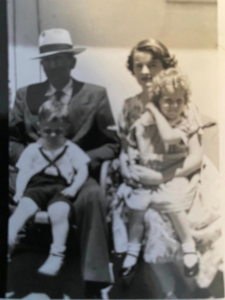 A vintage black-and-white family portrait featuring four individuals. On the left, a man in a suit and hat sits with a young boy on his lap. The boy wears a light-colored outfit with a dark cross strap and white socks. To the right, a woman elegantly dressed in a patterned blouse holds a little girl. The girl, wearing a dress with floral patterns and ruffled details, sits comfortably on her mother’s lap. The family poses together on a bench, displaying a formal yet affectionate demeanor. The background is softly blurred, focusing attention on the family.
