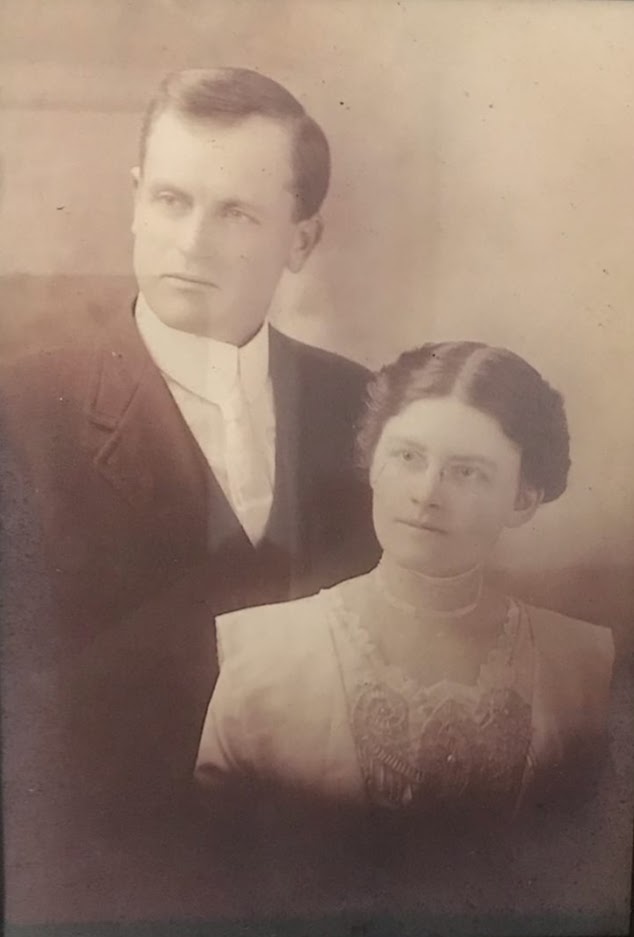 Vintage sepia-toned portrait of a couple, likely from the early 20th century. The man, presumably Olger, is dressed in a dark suit and tie with a solemn expression, looking slightly to the side. The woman, presumably Alma, is wearing a high-collared blouse adorned with intricate lace details, and her hair is styled back. She has a serene expression and is looking directly at the camera. They are positioned closely together, suggesting intimacy and partnership. The photograph has a soft focus, typical of the era, adding a timeless quality to their image.