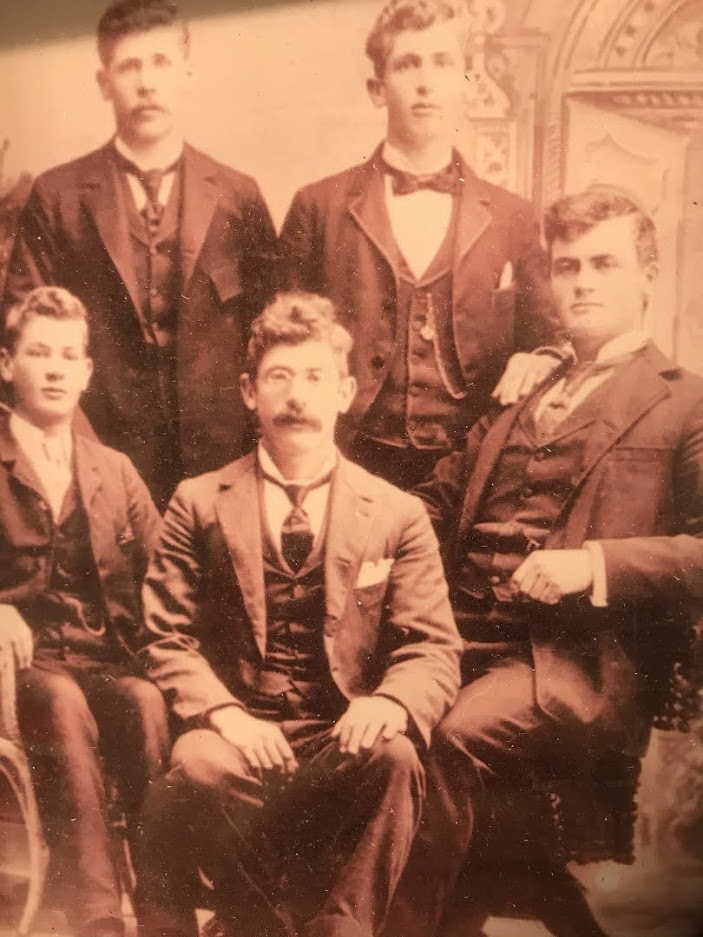Sepia-toned photograph of five men, identified as Herman (far right) and his brothers, dressed in formal late 19th or early 20th-century attire. The men wear dark suits with high collars, vests, and ties, seated and standing together in a studio setting with an ornate backdrop. Herman sits at the far right, exuding a confident expression, while his brothers display varying expressions of solemnity and composure, highlighting a moment of family unity and strength.