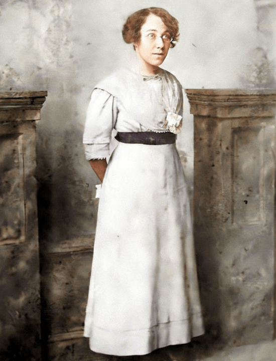 "A colorized portrait of Daisy standing in a formal pose, wearing a white dress with a dark sash at the waist. Her hair is styled softly, and she gazes slightly to the side. She stands between two ornate columns, against a backdrop that suggests a studio setting."