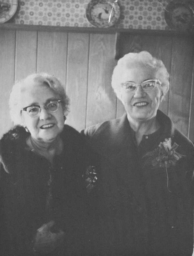 "A black-and-white photo of Daisy and Sylvia standing closely together, both smiling warmly. They are dressed in formal attire, one wearing a fur-lined coat and the other a corsage. A paneled wall and decorative plates are visible in the background. The women appear to share a deep bond."