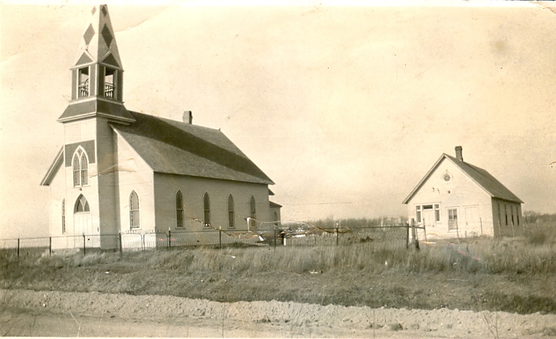 A historical photograph of Lost Island Lutheran Church, a wooden structure with a tall steeple and Gothic-style windows, set on a grassy landscape in rural Iowa. To the right of the church stands a smaller white building, likely used for church activities or schooling.