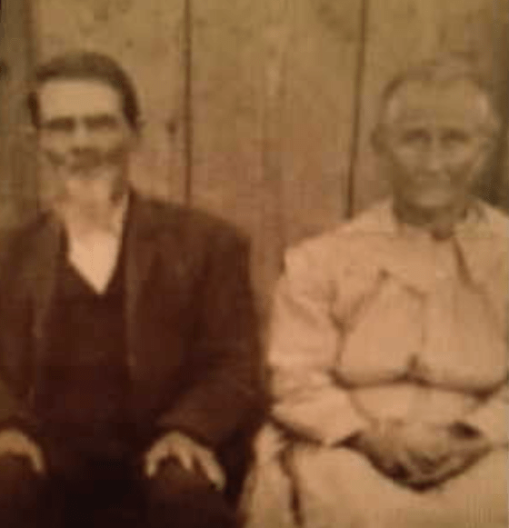 A sepia-toned historical photograph of an older couple seated side by side in front of a wooden wall. The man, identified as Daniel Pyatt, wears a dark suit with a white beard and mustache, while the woman, Mary Pyatt, wears a light-colored long-sleeve dress. Both have solemn but gentle expressions, reflecting their life together in the late 19th or early 20th century