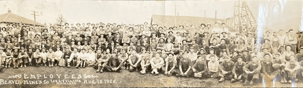 A large group photograph of the employees of Anna Beaver Mines in 1926. The workers, dressed in overalls and work attire, are posed outside near mining structures with a dog seated in the foreground.