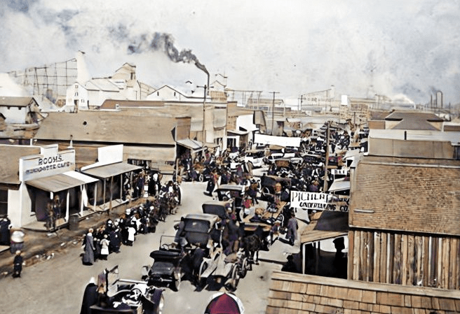 A bustling street scene in Picher, Oklahoma, during the 1920s, showing rows of buildings, a crowd of people, and smoke rising from industrial facilities in the background.