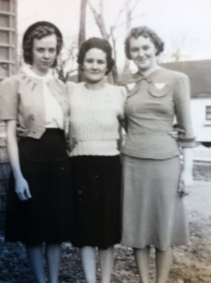 A black-and-white photograph of three women, Dorris, Lorraine, and Joyce Mitchell, standing together outdoors in daytime. They are dressed in skirts and blouses, with hairstyles reflecting mid-20th-century fashion. The background shows a house and leafless trees, suggesting late fall or early spring. They stand close together, smiling subtly at the camera.