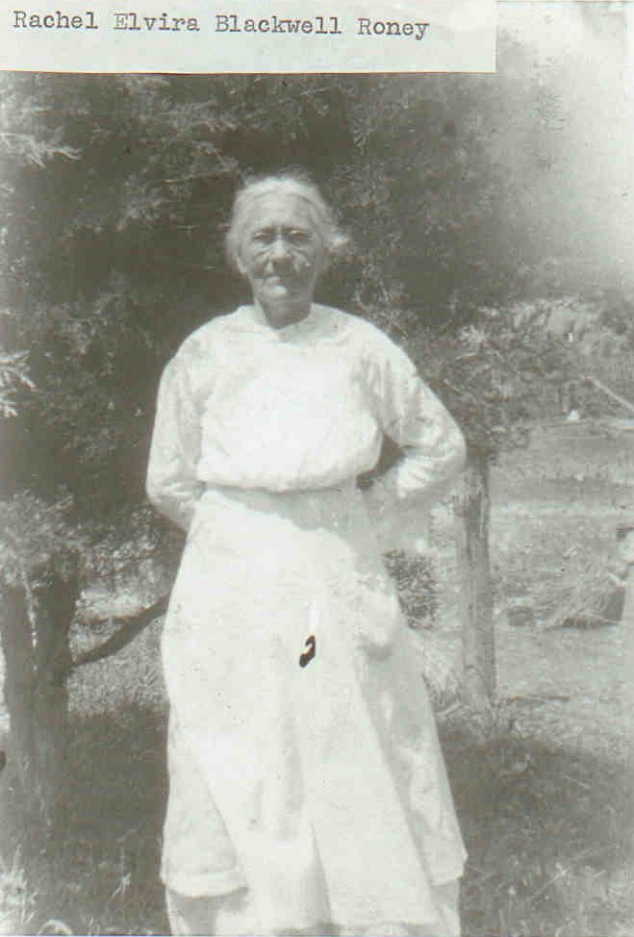A black-and-white portrait of Rachel Elvira Blackwell Roney, standing outdoors in front of leafy trees. She wears a light-colored, long-sleeved dress with an apron tied at the waist. Her hair is pulled back, and she has a calm, dignified expression on her face. A typewritten label at the top of the image identifies her by name.