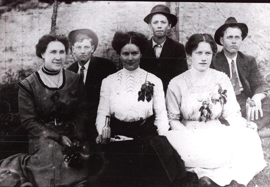 A black-and-white photo of six young people sitting and standing in two rows outdoors. In the front row, three women are seated, wearing light blouses with high collars and floral corsages pinned to their dresses. One of the women holds a bottle. Behind them stand three young men wearing dark jackets and hats. Their expressions are neutral to slightly serious, typical of early 20th-century portrait photography. The background features a faint outline of a fence.
