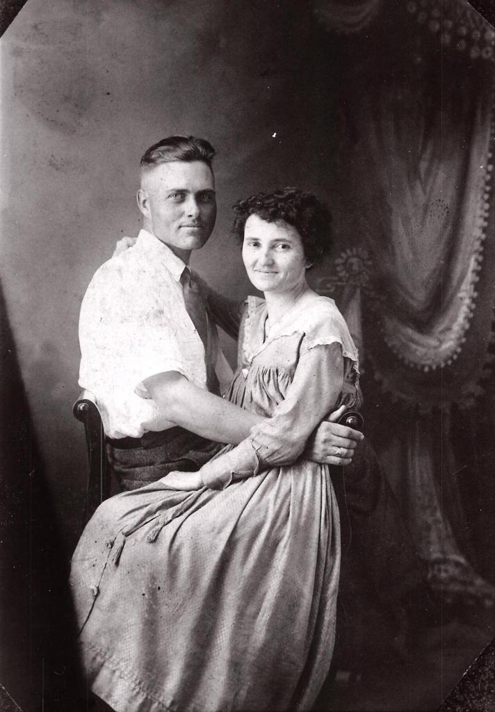 A black-and-white studio portrait of a young couple, Morris Roney and his fiancée Lilly. Morris sits in a chair with Lilly on his lap, both facing the camera. Morris wears a light-colored shirt and tie with his sleeves rolled up, and Lilly wears a light, flowing dress. They both have gentle smiles, and there’s a close, affectionate warmth in their posture. The backdrop features ornate drapery.