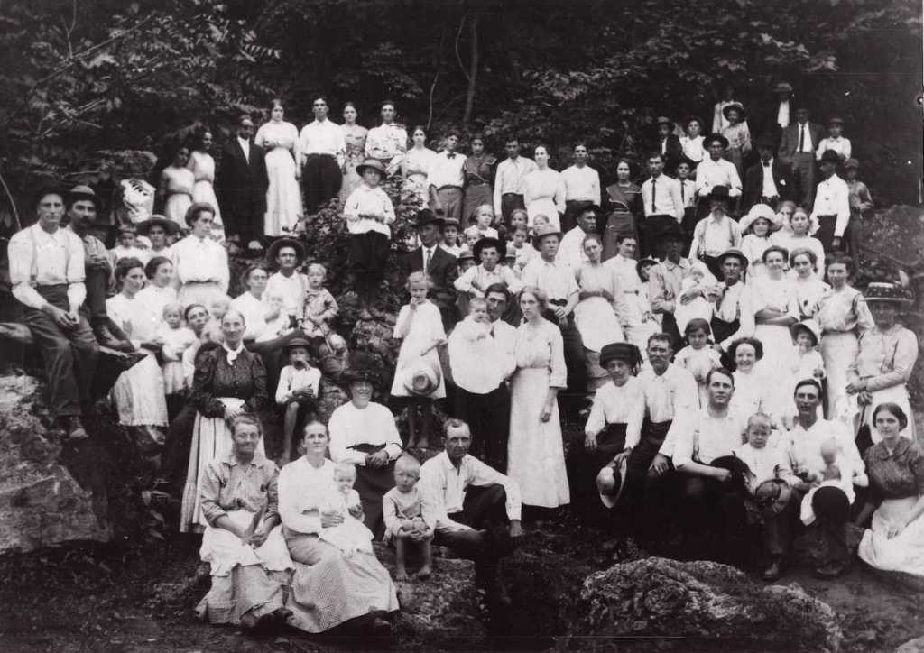 A large black-and-white photograph of an extended family gathered outdoors on rocky terrain, surrounded by trees. The group consists of men, women, and children of various ages, all dressed in early 20th-century attire. The women wear long dresses with high collars, and the men are in shirts, ties, and hats. Some people are seated on rocks while others stand in rows behind them, creating a layered effect.