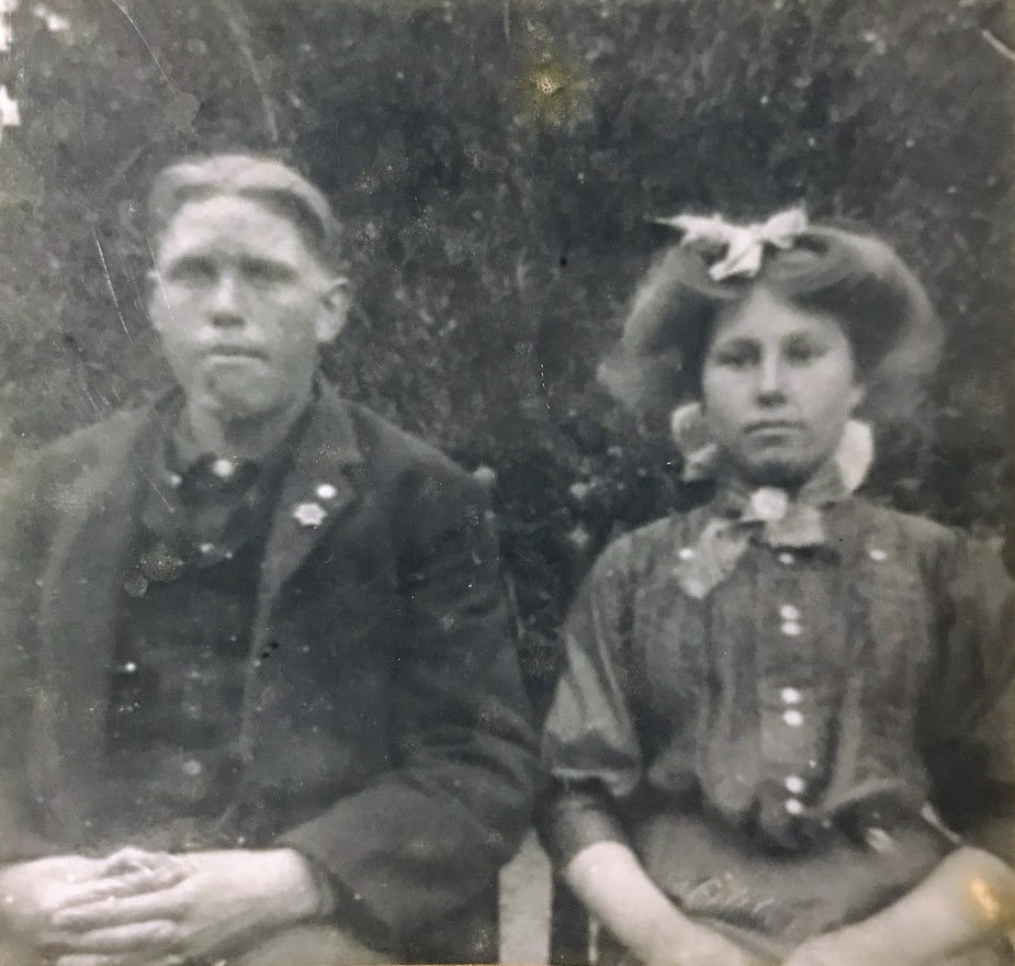 A black-and-white photograph of a young couple seated side by side. The man, dressed in a dark suit with a buttoned jacket, has neatly combed hair and a solemn expression. The woman wears a high-necked blouse with decorative buttons and a bow in her voluminous hair, her gaze steady and serious. The background is blurred, likely an outdoor setting, suggesting a formal portrait from the early 1900s.