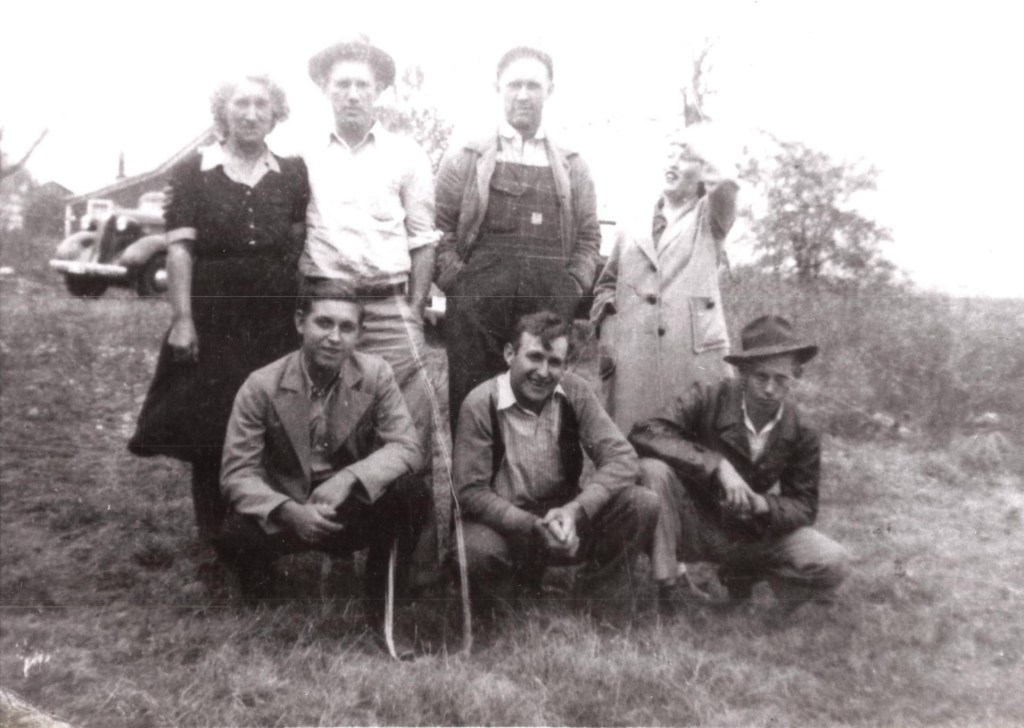 A black-and-white group photograph taken outdoors, featuring a mix of men and women, likely Myrtle’s children, dressed in overalls, button-down shirts, and jackets. Some are standing, while others crouch in the foreground. A vintage car and a farmhouse are visible in the background, setting the scene in a rural environment.