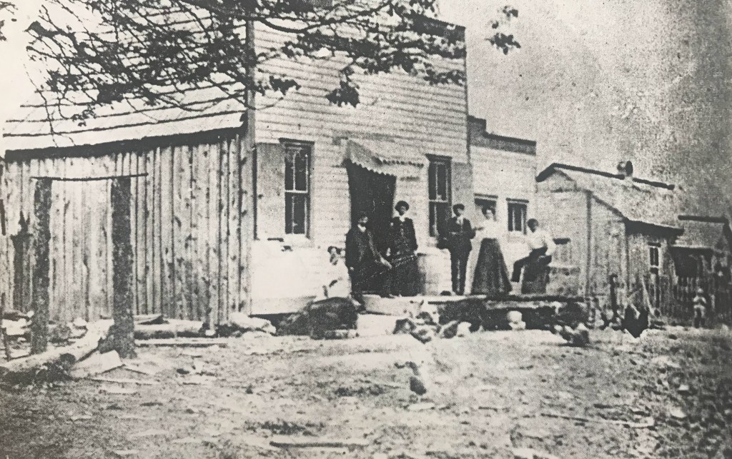 A grainy black-and-white image of a rustic wooden storefront, with a group of people standing on the porch. The building appears to serve as a general store and post office, evidenced by barrels and supplies outside. The individuals, dressed in period-appropriate attire, stand in formal yet relaxed postures, marking the importance of the establishment to their daily lives.